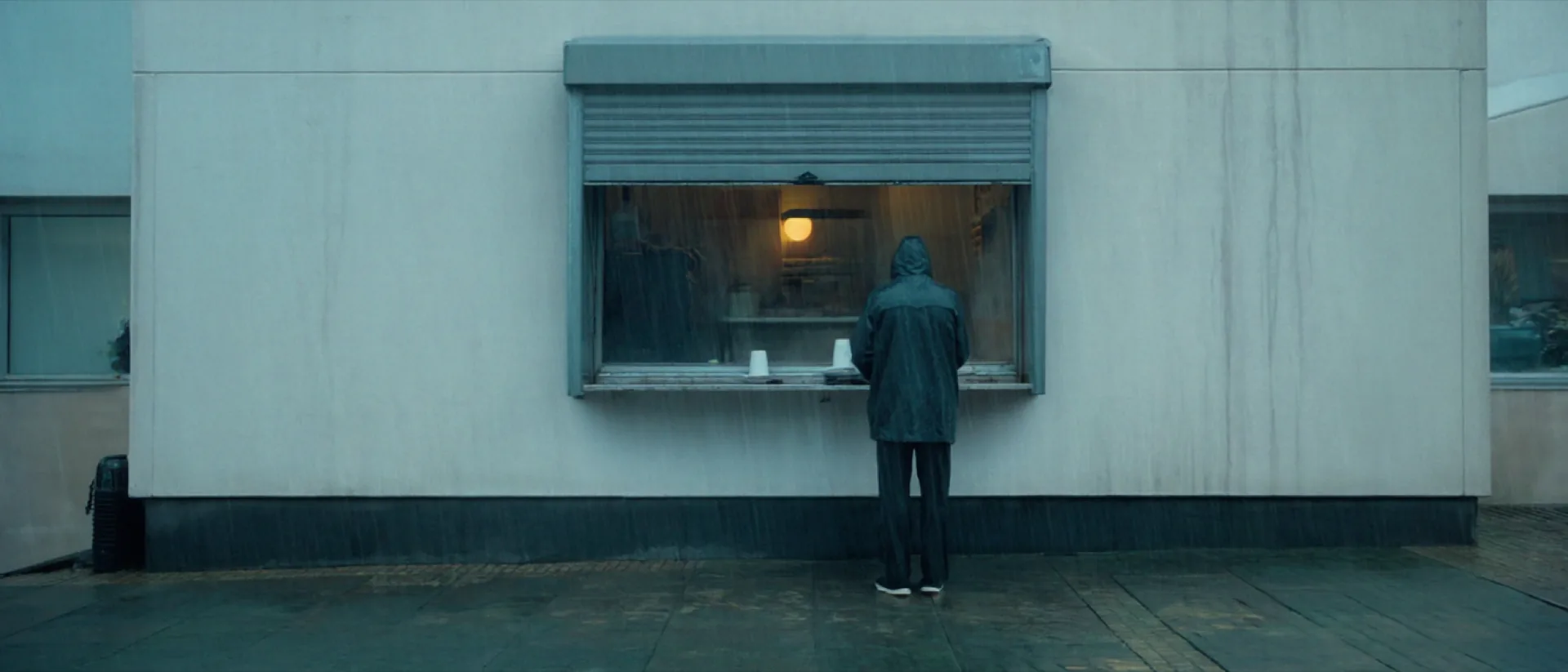 The open serving hatch of the Kaffeeklappe on a wet Winterhude street, two people in rain jackets waiting under the overhang