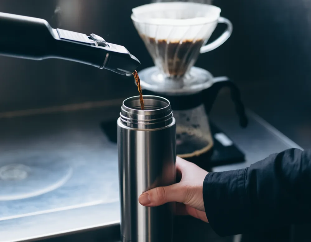A hand holding a dented steel vacuum flask under the brewer spout as dark coffee runs in